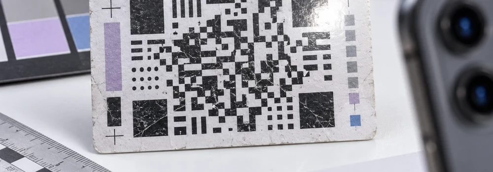 A worn abstract matrix code specimen under laboratory inspection, with phone camera lenses softly out of focus.